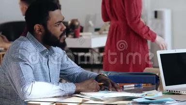 快乐的年轻非洲男人，穿着正式的衣服，坐在繁忙的现代办公桌上接受工作面试。 健康的工作场所。